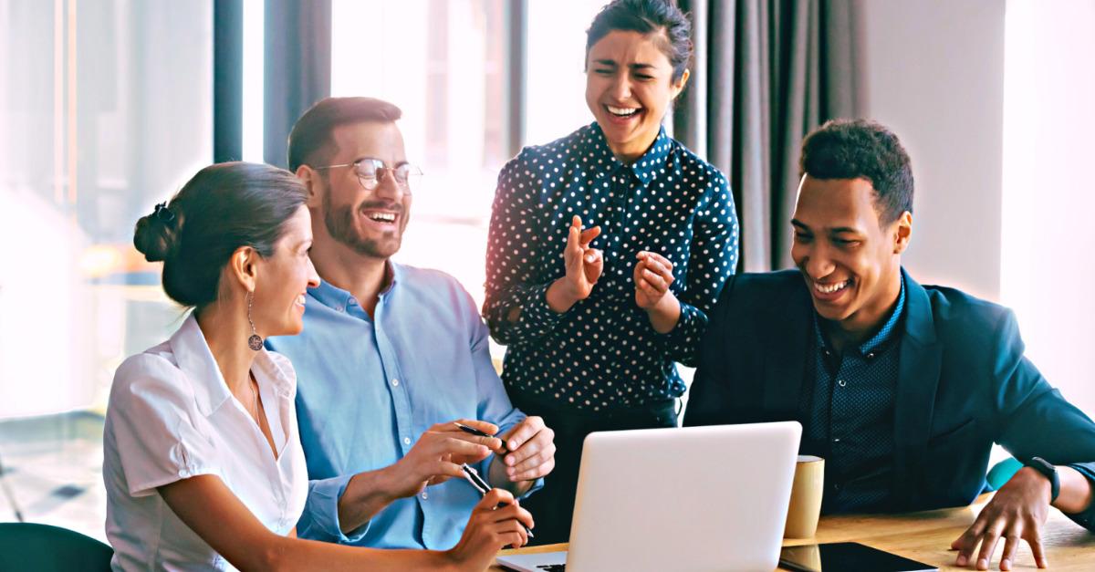 Pessoas sentadas em uma mesa de reuniões, em ambiente profissional diante de um computador. Elas estão rindo e felizes, demonstrando que colaboradores descansados ficam mais motivados.