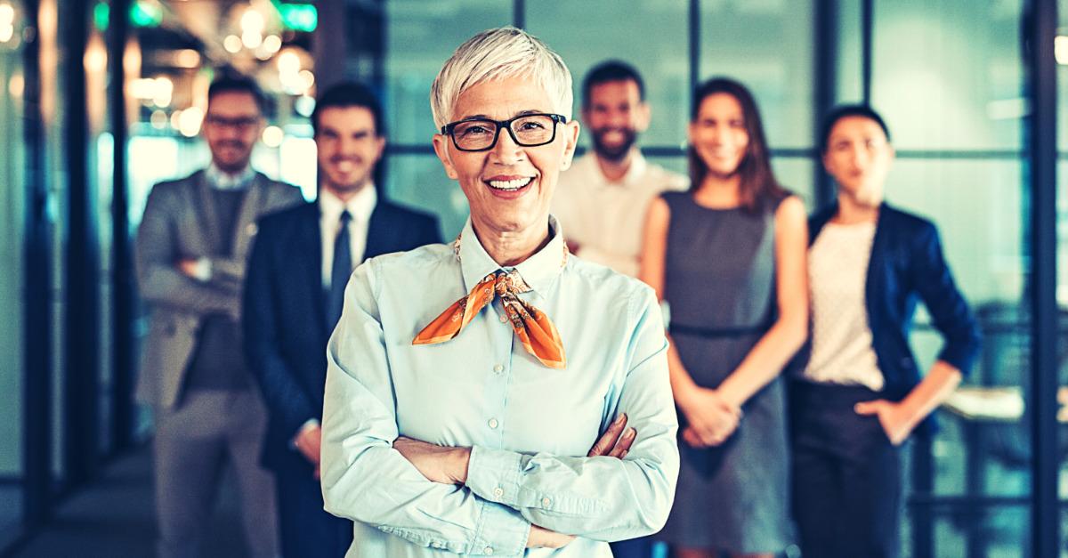 Mulher líder a frente, com os braços cruzados e um sorriso. Atrás dela estão uma equipe de 5 pessoas sorridentes. 