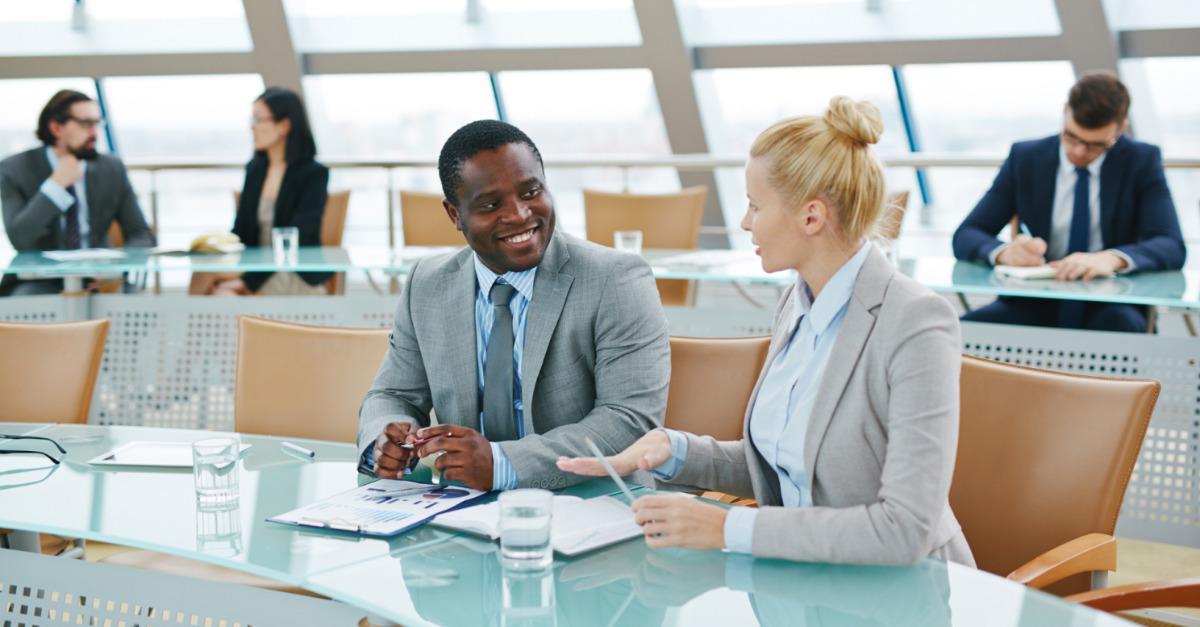 Homem e mulher vestindo trajes sociais e em um ambiente de trabalho. Estão sentados um ao lado do outro, sorrindo e conversando. A imagem simboliza o poder da comunicação positiva nos ambientes de trabalho. 