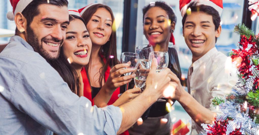 Pessoas com toucas de natal em ambiente decorado. Elas estão sorrindo e brindando em jum ambiente aparentemente corporativo.